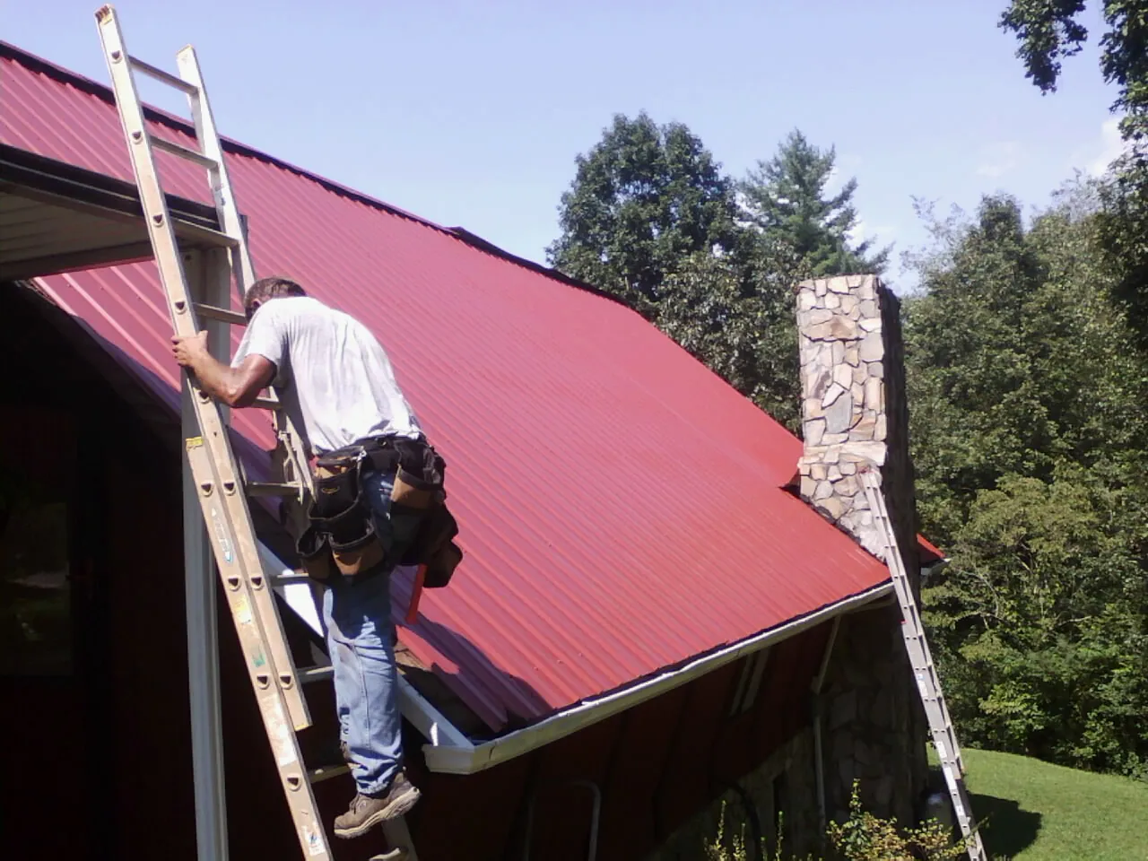 Roofer on a ladder beside a red metal roof and stone chimney — Soffit & Fascia Repair by Roofing Brothers in South Hutchinson